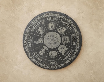May include: A circular dark gray slate plaque featuring an engraved wheel of the year. The design includes a central pentagram surrounded by eight segments labeled Ostara, Beltane, Litha, Lammas, Mabon, Samhain, Yule, and Imbolc with dates.