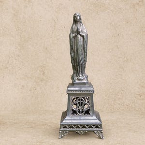 May include: A silver-colored statue of the Virgin Mary, standing on a decorative pedestal. The statue features detailed robes and a rosary. The pedestal has an ornate, openwork design.