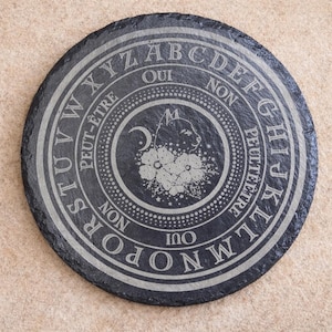 May include: A circular, dark grey slate coaster with etched white lettering and designs. The design includes the alphabet, the words "Oui" and "Non", and a central image of a cat, moon, and flowers. The coaster is on a light brown surface.