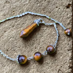 May include: A necklace with a tiger's eye pendant and beads. The pendant is a brown, elongated crystal shape, and the necklace has three round beads. The chain is silver-coloured.