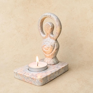 Peut inclure: Un bougeoir en pierre marbrée de couleur claire, composé d'une base rectangulaire et d'une silhouette verticale abstraite avec un sommet circulaire et un motif en spirale sur le torse. Une petite bougie chauffe-plat blanche est allumée.