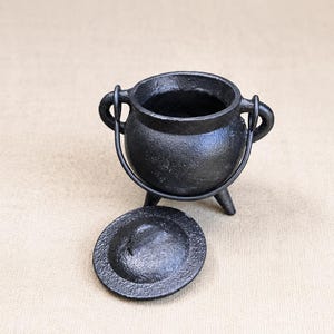 Puede incluir: Un pequeño caldero de hierro fundido negro con cuerpo redondeado, tres patas cortas y dos asas laterales. Una tapa circular a juego de hierro fundido negro está colocada frente al caldero. La superficie del metal tiene un acabado mate texturizado.