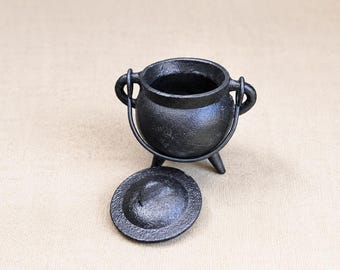 May include: A small black cast iron cauldron with a rounded body, three short legs, and two side handles. A matching circular black cast iron lid is positioned in front of the cauldron. The surface of the metal has a textured, matte finish.