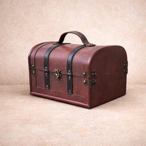 May include: A small, antique-style wooden chest with a curved top and a leather handle. The chest is stained a dark reddish-brown colour and features black leather straps and metal hardware. The chest measures approximately 30 cm long, 20 cm wide, and 20 cm high.