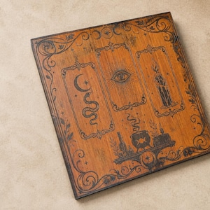 Op de afbeelding: Een rechthoekig houten bord met een donkerbruine afwerking en zwarte, in laser gegraveerde illustraties. Het ontwerp bevat drie verticale rechthoekige kaders met een slang met een maansikkel, een oog, een kaars en een rokende ketel.