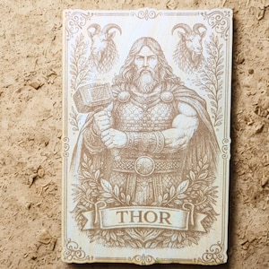 Könnte beinhalten: Holztafel mit einer Sepia-Gravur von Thor, der einen Hammer hält. Das Bild zeigt detaillierte Darstellungen seiner Rüstung, seines Bartes und das Wort "THOR" auf einem Banner. Zwei Widder und Blätter umrahmen die zentrale Figur und bilden einen dekorativen Rahmen.