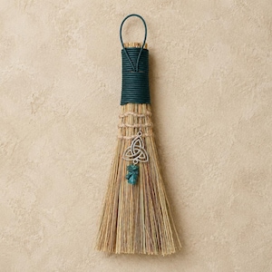 May include: A small hand broom made of natural tan straw fibres with a dark teal wrapped handle and a hanging loop. The broom is decorated with a silver-toned metal triquetra charm and a small, rough-cut teal stone pendant attached by twine.