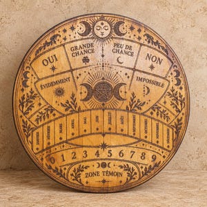 May include: A circular wooden board featuring intricate pyrography designs including a central pentagram, moon phases, numbers zero through nine, months of the year, and French text such as OUI, NON, GRANDE CHANCE, PEU DE CHANCE, and ZONE TÉMOIN.