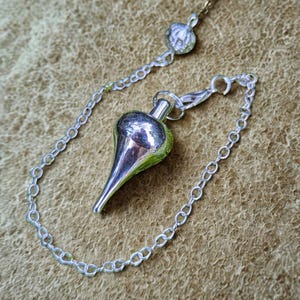 Op de afbeelding: Een zilverkleurige pendel met een druppelvormig lichaam en een puntige punt, verbonden met een ketting. De ketting is ook zilverkleurig en heeft een klein decoratief element aan de bovenkant. De achtergrond is een getextureerd, bruin oppervlak.