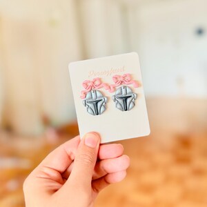 May include: Silver Mandalorian helmet earrings with black detailing and pink bows, displayed on a cream card. The earrings are held by a hand. The card has the text "DreamyJewel".
