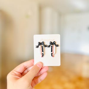 Pode incluir: Um par de brincos com uma fita preta, um corpo branco e detalhes vermelhos e pretos. Os brincos são exibidos em um cartão branco com o texto "PennyJewel".
