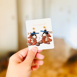 May include: Novelty earrings with a cartoon dog design. The earrings are brown, black, and white with blue accents. They are attached to a white card with the text "Desingjewel".