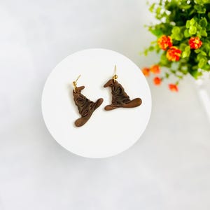 May include: A pair of brown witch hat earrings with gold-colored hooks. The earrings are on a white circular surface, with a blurred background of green leaves and orange flowers. The earrings are a whimsical accessory.