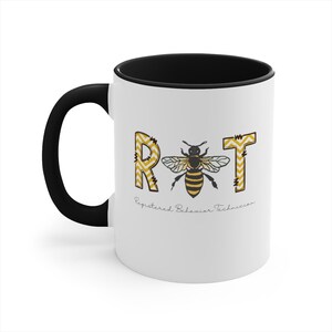 May include: White ceramic mug with a black handle and interior. The mug features the letters "RBT" in yellow and white chevron pattern, with a bee illustration. The text "Registered Behavior Technician" is below the letters.