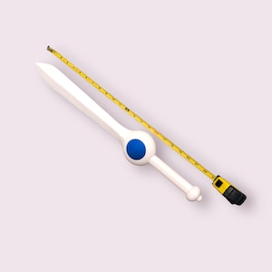 May include: A white plastic sword with a blue circle on the handle. The sword is lying on a yellow measuring tape.