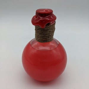 Può includere: Una piccola bottiglia rotonda di vetro con un sigillo di cera rossa e un tappo di spago marrone. La bottiglia è piena di un liquido rosso brillante.