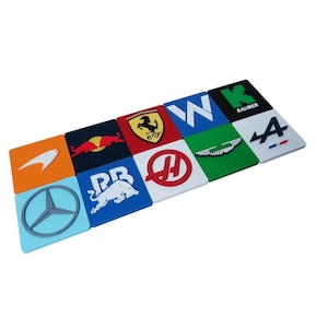 May include: A collection of square coasters featuring Formula 1 team logos. The coasters display logos for McLaren, Red Bull, Ferrari, Williams, Sauber, Aston Martin, and others. Each coaster is a different color.