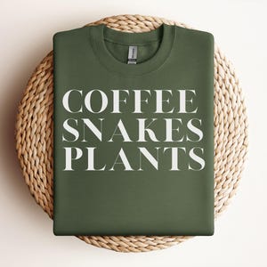 Może przedstawiać: Oliwkowo-zielona bluza z napisem "COFFEE SNAKES PLANTS" dużymi, białymi, wielkimi literami. Bluza jest prezentowana na okrągłej, tkanej podkładce z wikliny.