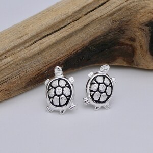 May include: A pair of silver turtle stud earrings. The turtles are detailed with a textured shell and have a black outline.