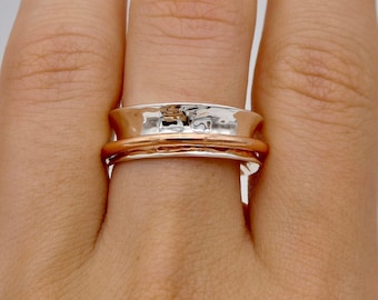 May include: A silver and rose gold ring with a textured design. The ring features a wide silver band with a hammered texture and a rose gold band. The ring is shown on a finger.