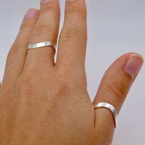 May include: Two silver rings are worn on a hand. The rings are simple, flat bands with a slightly curved design. The rings are made of a shiny, reflective metal, and are set against a plain background.
