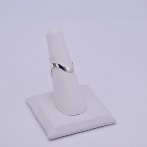 May include: A silver-colored ring with a V-shaped design, displayed on a white ring holder. The ring has a smooth, polished surface and a minimalist aesthetic. The ring is a piece of jewelry.