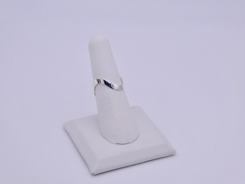 May include: A silver-colored ring with a V-shaped design, displayed on a white ring holder. The ring has a smooth, polished surface and a minimalist aesthetic. The ring is a piece of jewelry.