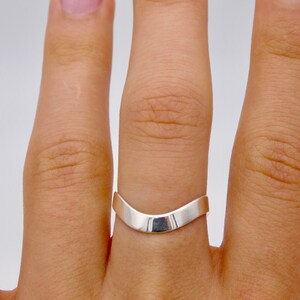 May include: A silver-colored ring with a smooth, curved design. The ring has a V-shape, with a polished, reflective surface. The ring is worn on a finger, showcasing its minimalist and modern aesthetic.