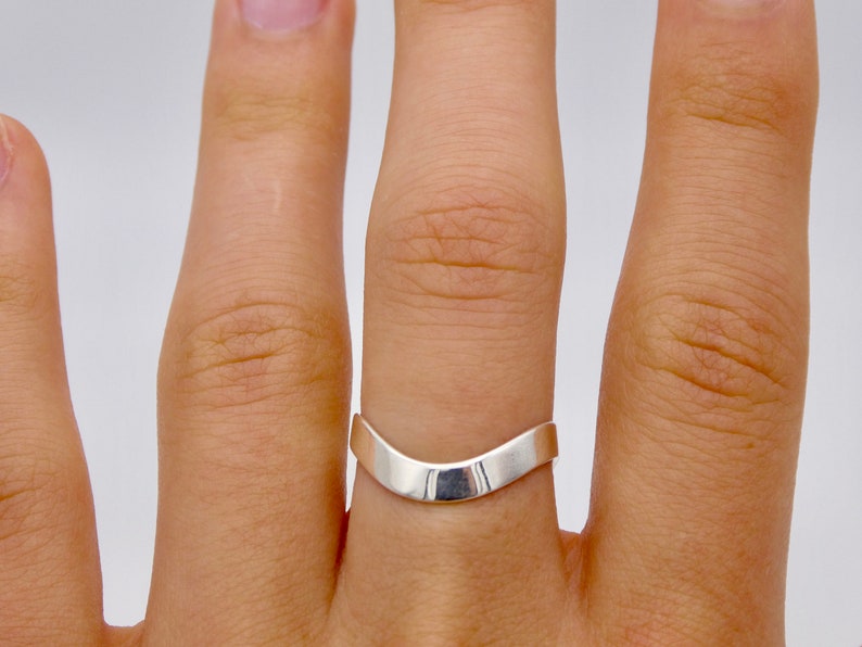 May include: A silver-colored ring with a smooth, curved design. The ring has a V-shape, with a polished, reflective surface. The ring is worn on a finger, showcasing its minimalist and modern aesthetic.