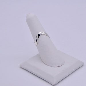 May include: A polished silver ring with a smooth, curved design. The ring is displayed on a white, angled stand, showcasing its simple elegance. The ring's surface reflects light, highlighting its metallic sheen. The ring is a minimalist piece of jewelry.