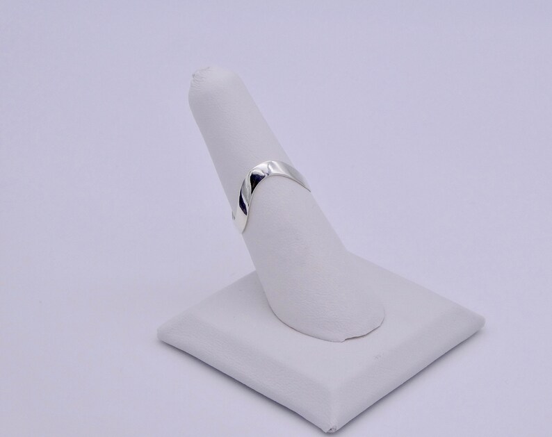 May include: A polished silver ring with a smooth, curved design. The ring is displayed on a white, angled stand, showcasing its simple elegance. The ring's surface reflects light, highlighting its metallic sheen. The ring is a minimalist piece of jewelry.