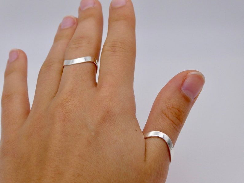 May include: Two silver rings are worn on a hand. The rings are simple, flat bands with a slightly curved design. The rings are made of a shiny, reflective metal, and are set against a plain background.