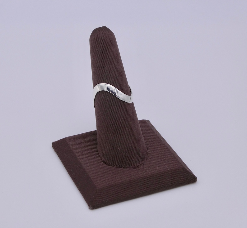 May include: A silver-colored ring with a wavy design. The ring is displayed on a brown ring holder. The ring's design is a simple, elegant wave shape. The ring is made of a shiny, reflective metal.