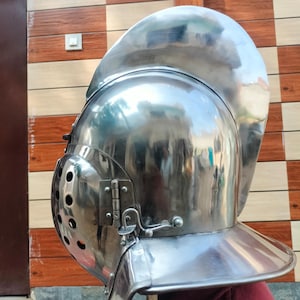 Puede incluir: Un yelmo medieval pulido, de color plateado, con la parte superior redondeada y una visera con múltiples agujeros pequeños. El yelmo tiene una visera con bisagras y un protector de cuello.