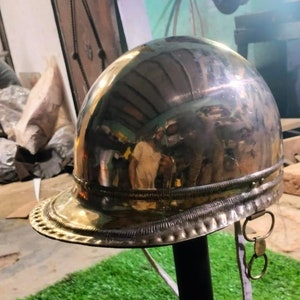 Könnte beinhalten: Ein polierter Metallhelm mit abgerundeter Krone und Krempe, präsentiert auf einem schwarzen Ständer. Der Helm spiegelt die Umgebung wider. Der Ständer steht auf einer quadratischen Basis vor Kunstrasen.