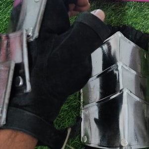 Puede incluir: Un par de guantes de cuero negro con placas de metal en la parte posterior de la mano y el antebrazo. Los guantes se llevan sobre un guante negro. Los guantes están sobre una superficie de hierba verde.
