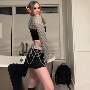 May include: A person wearing a black skirt and a silver chainmail top with long sleeves. The person is standing in a bathroom with a white bathtub and a black backpack on the floor.