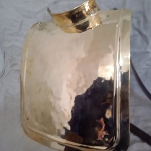 May include: A gold-colored metal breastplate with a rounded top and a smooth, slightly textured surface.