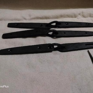 Peut inclure: Trois charnières en métal noir avec une boucle à une extrémité et un crochet à l'autre. Les charnières sont disposées sur une surface en tissu blanc. Il y a aussi un petit crochet en métal noir sur la surface.