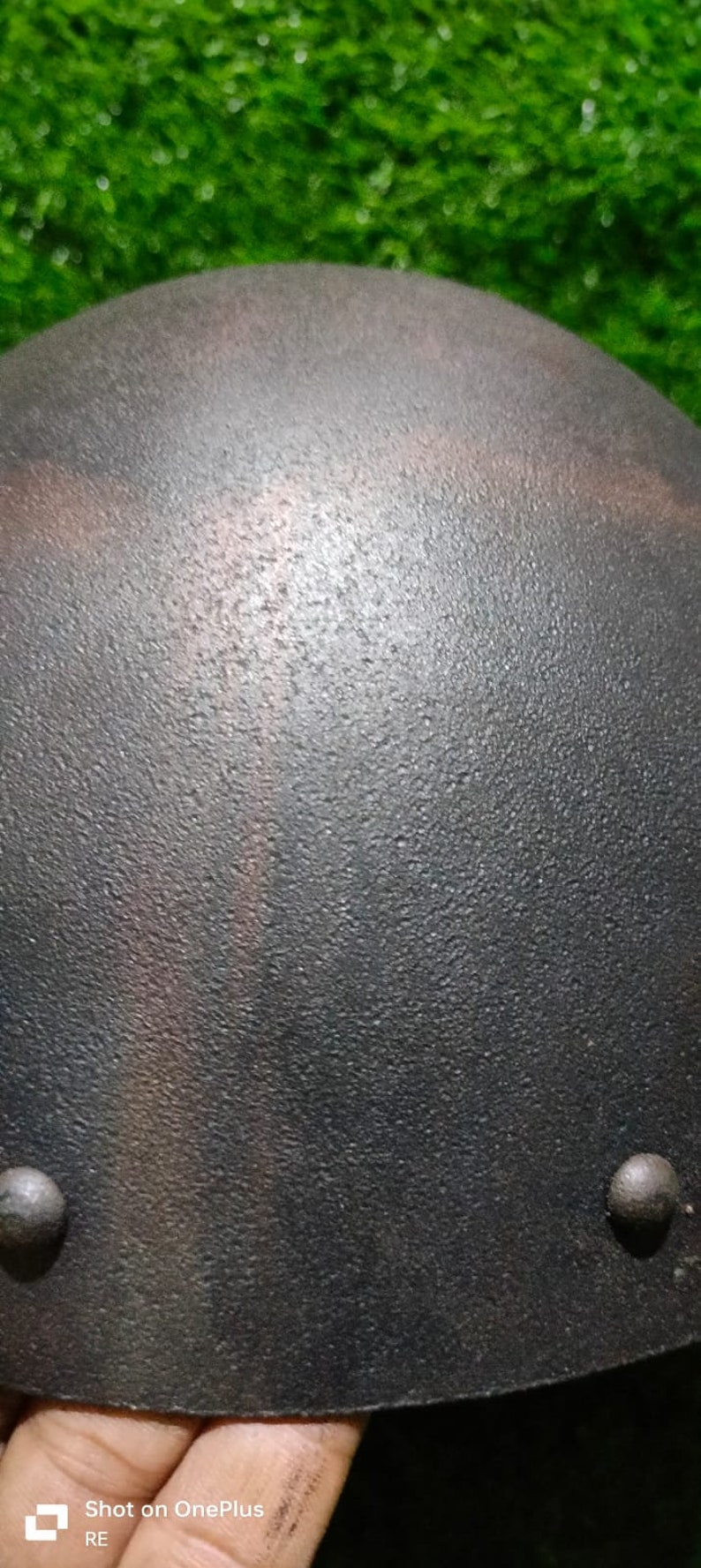 May include: A dark grey, textured, dome-shaped helmet with two metal rivets. The helmet has a subtle, iridescent sheen.