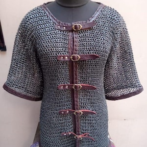 Puede incluir: Una camisa de cota de malla plateada con correas y hebillas de cuero marrón. La camisa tiene mangas cortas y un escote redondo.