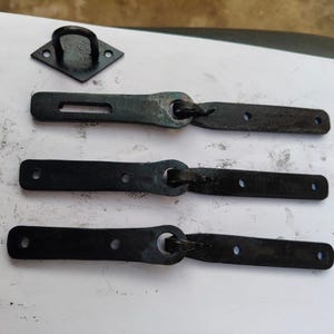 Könnte beinhalten: Drei schwarze Metall-Türverschlüsse mit einem Haken an einem Ende und einer Schlaufe am anderen. Die Verschlüsse sind in einer Reihe auf einer weißen Oberfläche angeordnet.