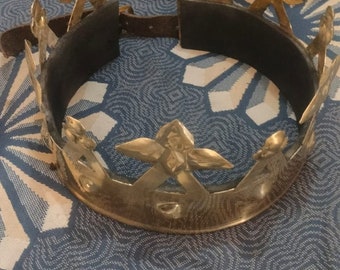 Puede incluir: Una corona dorada con una banda de cuero negro. La corona tiene un diseño en forma de estrella con cinco puntas en cada estrella.