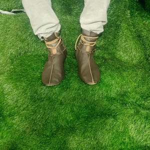 May include: Brown leather ankle boots with lace-up detailing. The boots are worn with light grey trousers, and the image is taken from a top-down perspective. The boots are on a bed of green artificial grass.