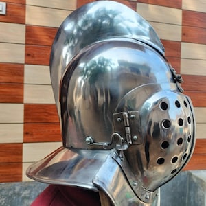 May include: A shiny, silver-coloured medieval helmet with a visor featuring multiple small holes. The helmet has a rounded top and a flared neck guard. The image shows the side view of the helmet, highlighting its intricate design and reflective surface.