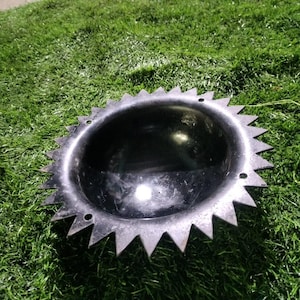 Könnte beinhalten: Eine schwarze Metallscheibe mit einem scharfen, gezackten Rand. Die Scheibe hat eine konkave Mitte und liegt auf einer grünen Grasfläche.