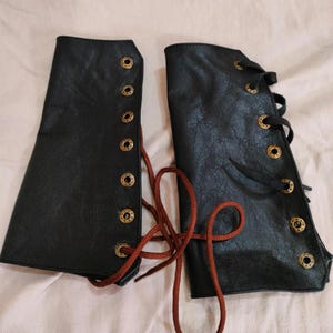 May include: A pair of black leather arm warmers with brown laces and gold metal eyelets. The arm warmers are tied together with the laces.