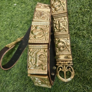May include: A gold-colored leather belt with a decorative buckle. The belt is adorned with square-shaped metal plates that have intricate designs.