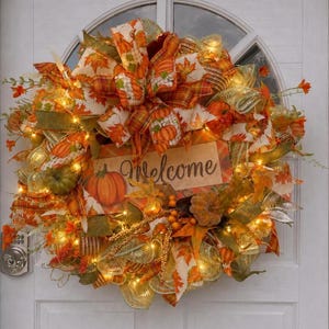 May include: A decorative autumn wreath featuring orange and green mesh, fabric ribbons with pumpkin and leaf patterns, small artificial pumpkins, and warm white string lights. A central rectangular sign displays the word Welcome in script.