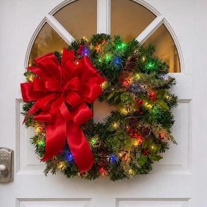 May include: A circular green artificial pine wreath featuring a large, vibrant red satin bow on the left side. The wreath is decorated with brown pinecones, clusters of red berries, and multicolored LED string lights glowing throughout.